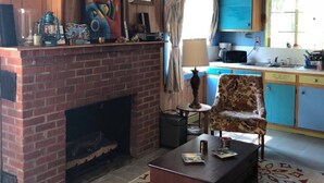 Een woonkamer met een open haard van baksteen, een kleurrijk abstract schilderij, een kamerbreed tapijt en een eethoek met blauwe kasten en een raam.
