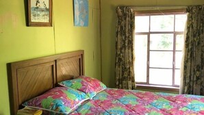 Een slaapkamer met een houten bed, bloemenbeddengoed, een nachtkastje met boeken, een raam met gordijnen en een muur met twee ingelijste foto’s.