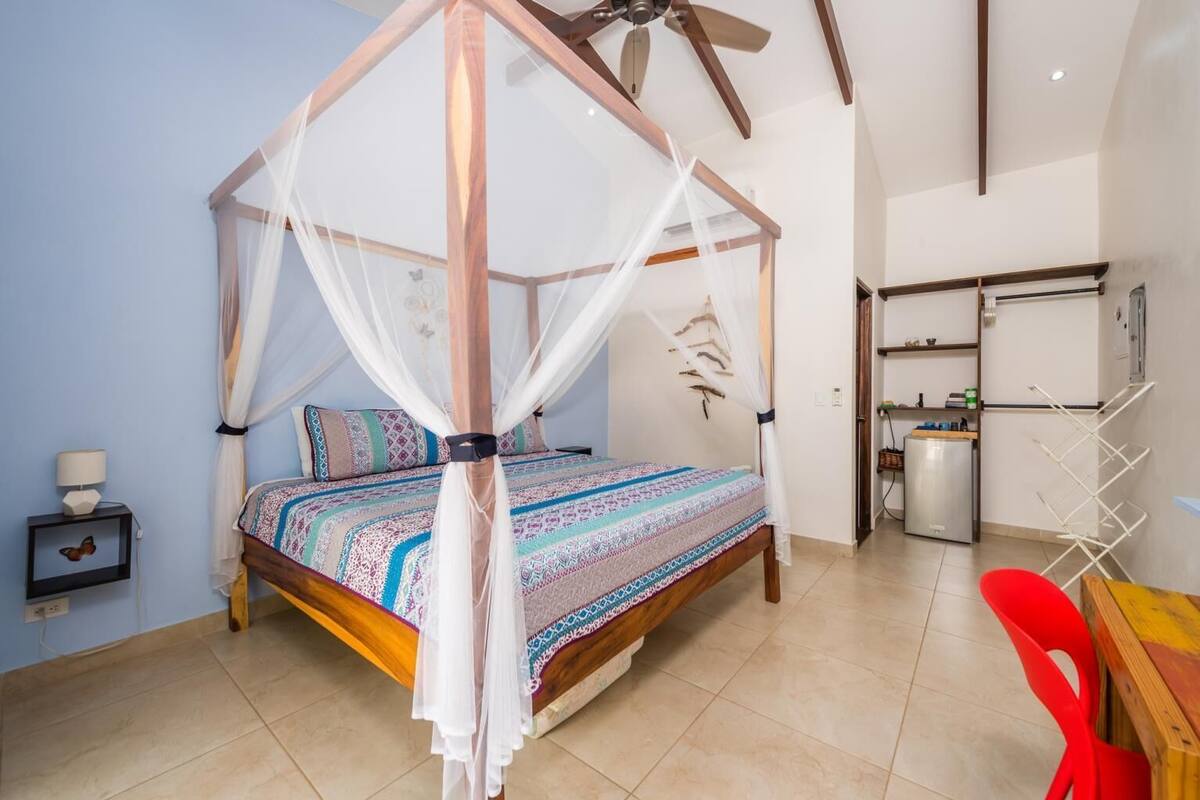 Une chambre à coucher avec un lit à baldaquin, une petite table de chevet avec une lampe, et un coin cuisine équipé d’un réfrigérateur et d’un évier.