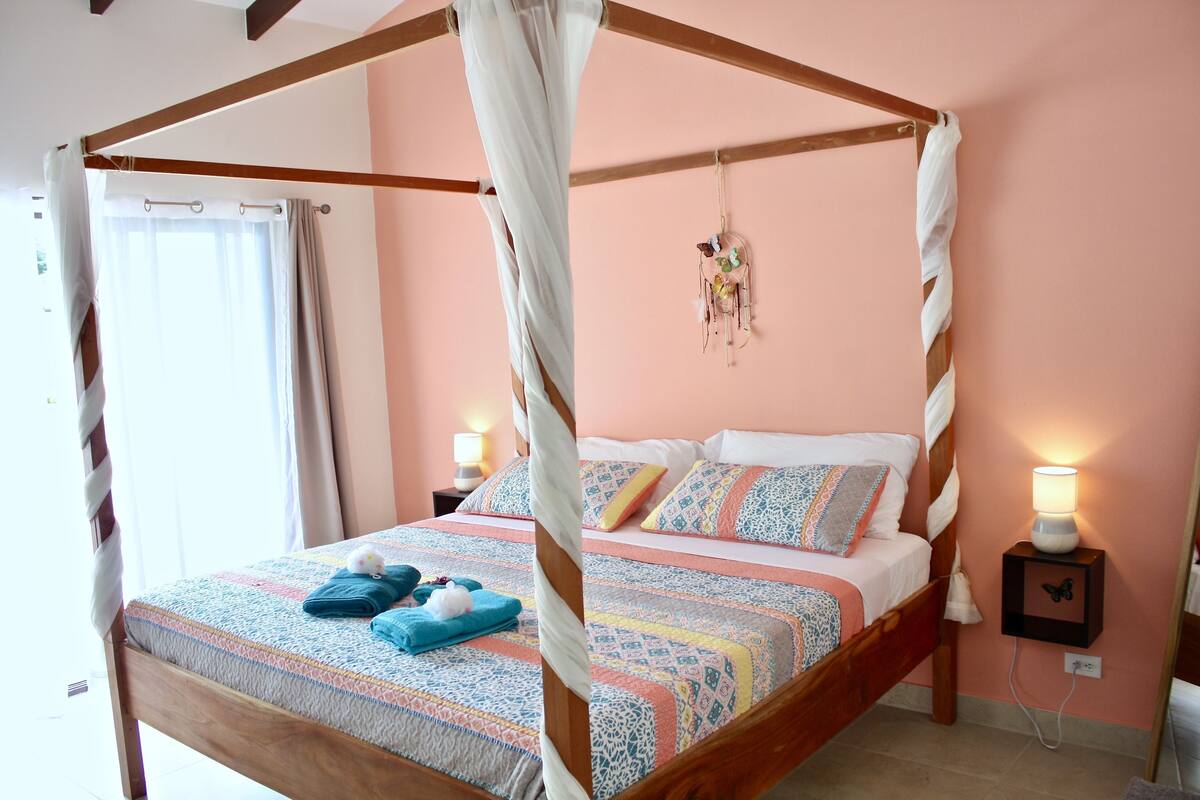 Un lit à baldaquin, une table de chevet avec une lampe et un attrape-rêves fixé au mur.