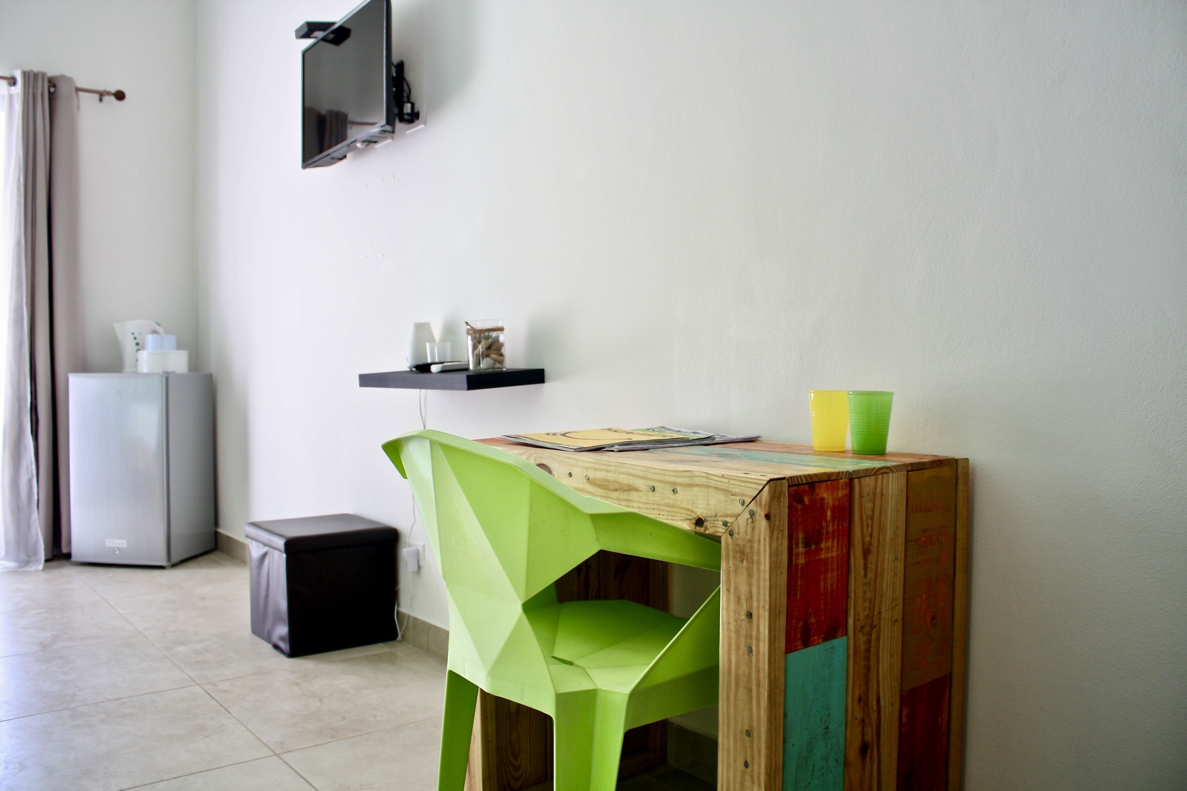 Una habitación con un escritorio de madera, una silla verde, un cubo de basura negro pequeño y un refrigerador gris, además de una estantería con objetos.