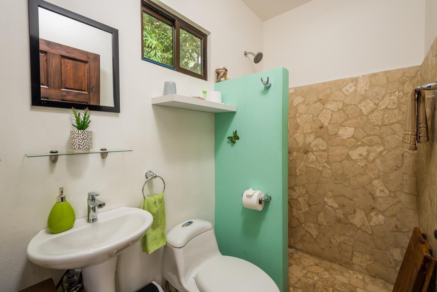 Une salle de bain avec un lavabo blanc, des toilettes et une douche. Il y a une fenêtre au-dessus de la douche, une serviette accrochée au mur et une petite étagère avec des articles de toilette.