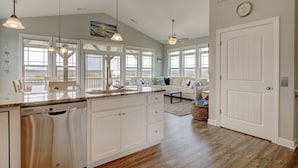A spacious kitchen with a large island, white cabinetry, and a dishwasher. There is a dining area with a white table and chairs, and a sofa with a view of the ocean. -  Across the street from the beach!!  4/3 with Spectacular views & ELEVATOR! (North Topsail Beach)