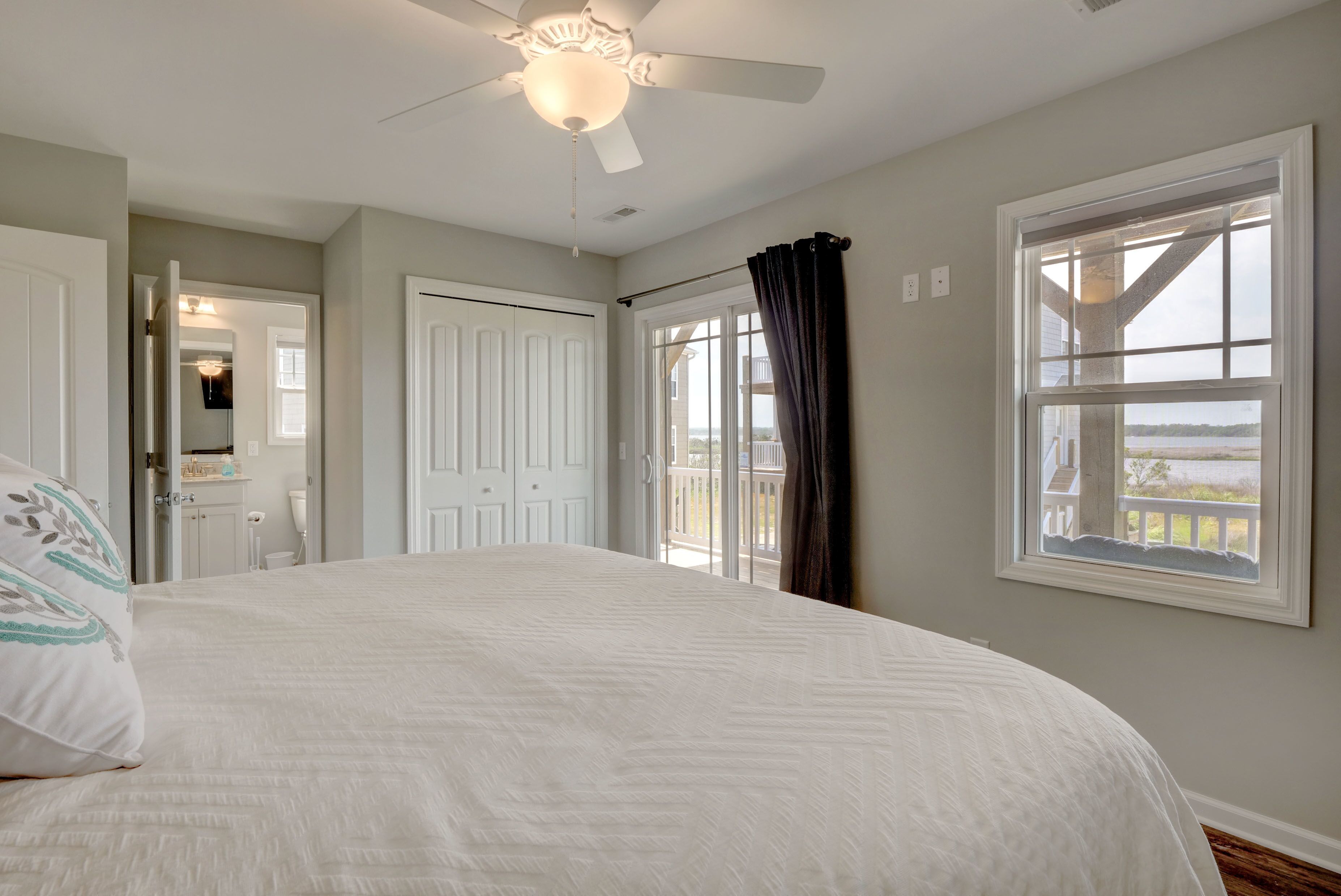 A bedroom with a large bed, a ceiling fan, a window with curtains, and a view of the outdoors.