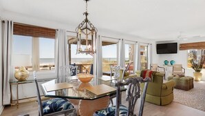 A spacious living room with a glass-top dining table, ornate chairs, a green sofa, and a large window offering a view of the beach. - Discounted rates! The Edge of the Edge of America! (Folly Beach)