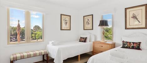 A bedroom with two beds, a bench, a nightstand, framed pictures, and a window with a view of greenery.