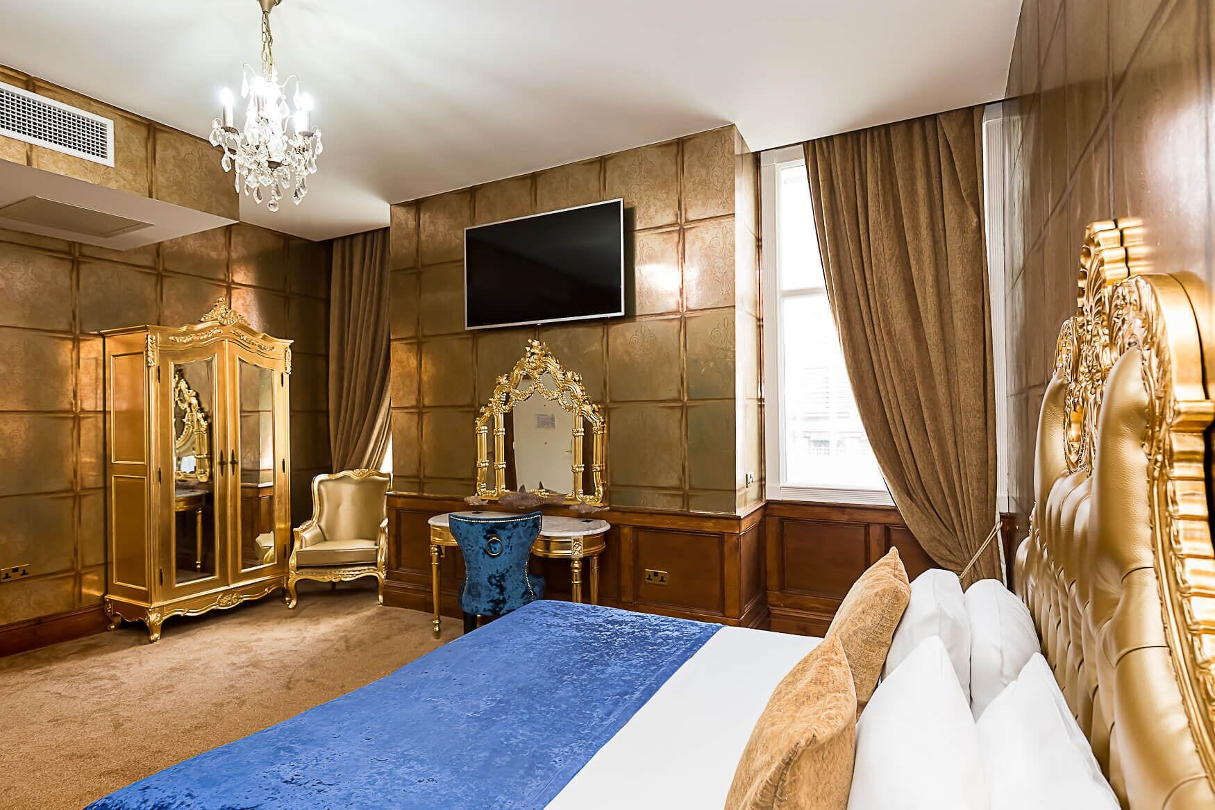 A luxurious bedroom with a large bed, a gold wardrobe, a vanity with a mirror, and a chandelier.