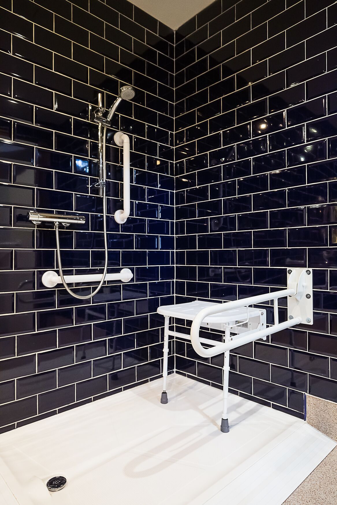 Un baño con paredes de azulejos negros, un asiento de ducha blanco y un cabezal de ducha manual.