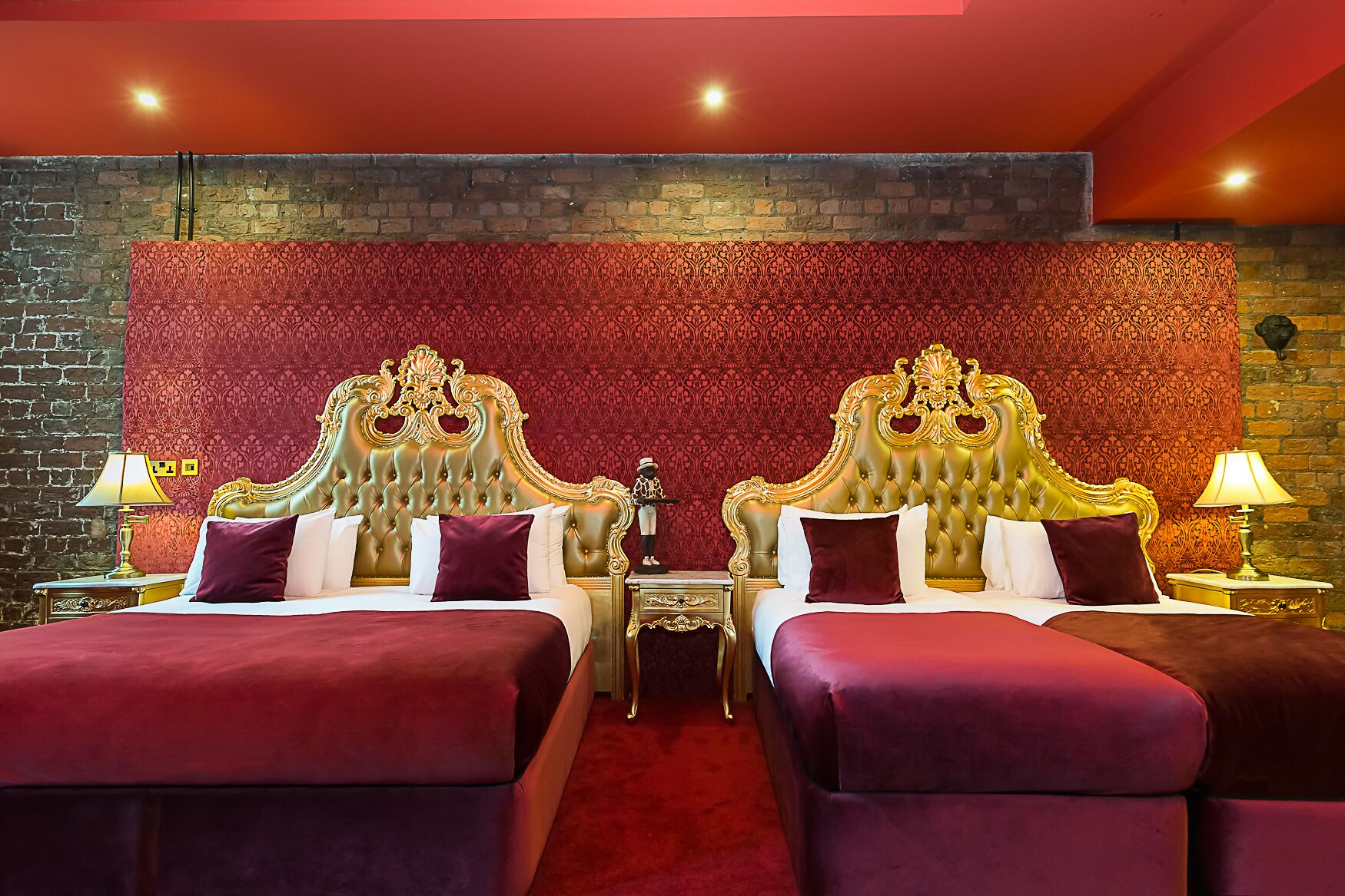 A luxurious hotel room with two beds, each with a gold headboard, red and white bedding, and a red carpet.