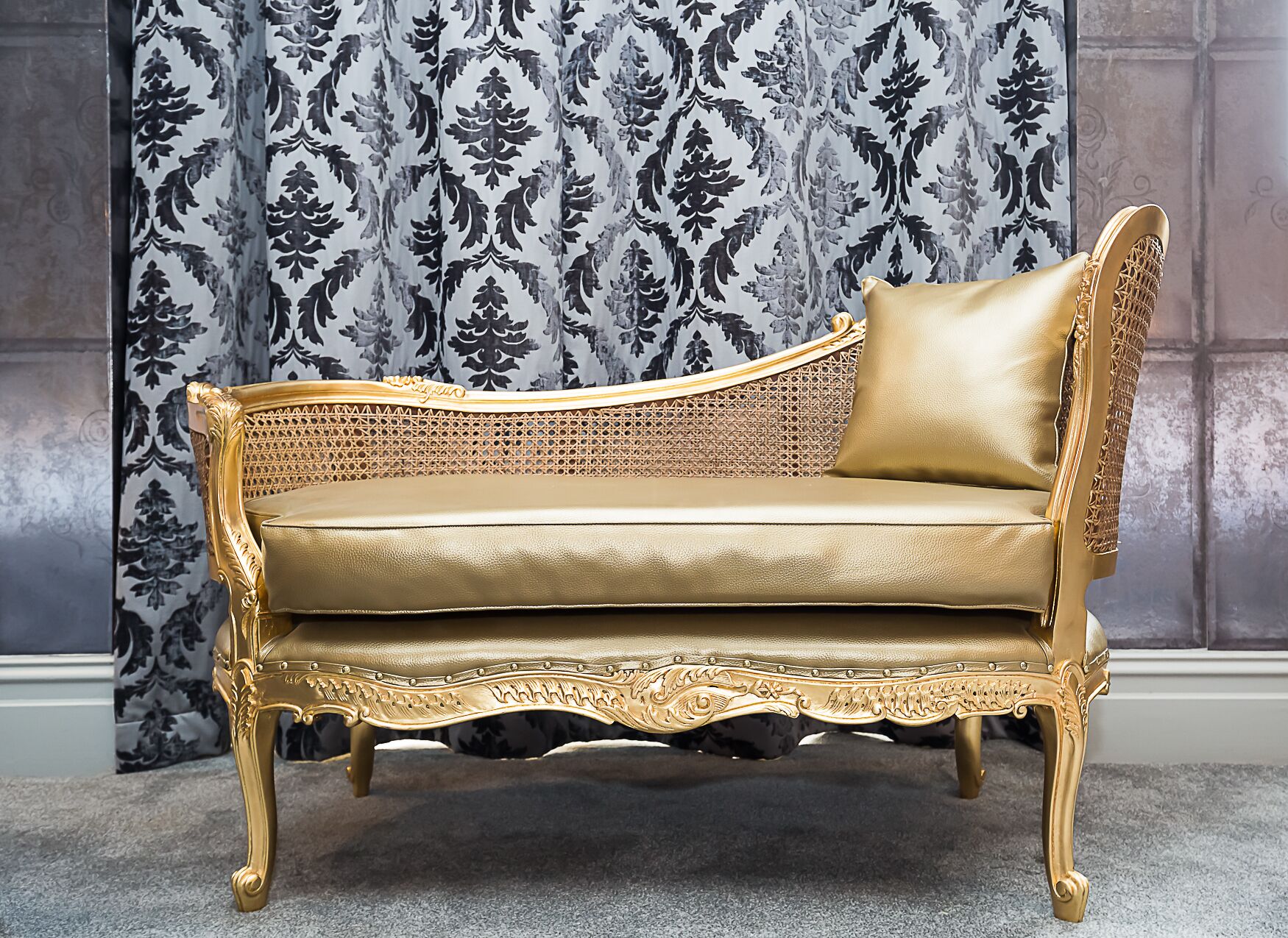 En gylden polstret sofa med detaljeret udskæring, placeret foran tapetbaggrund.