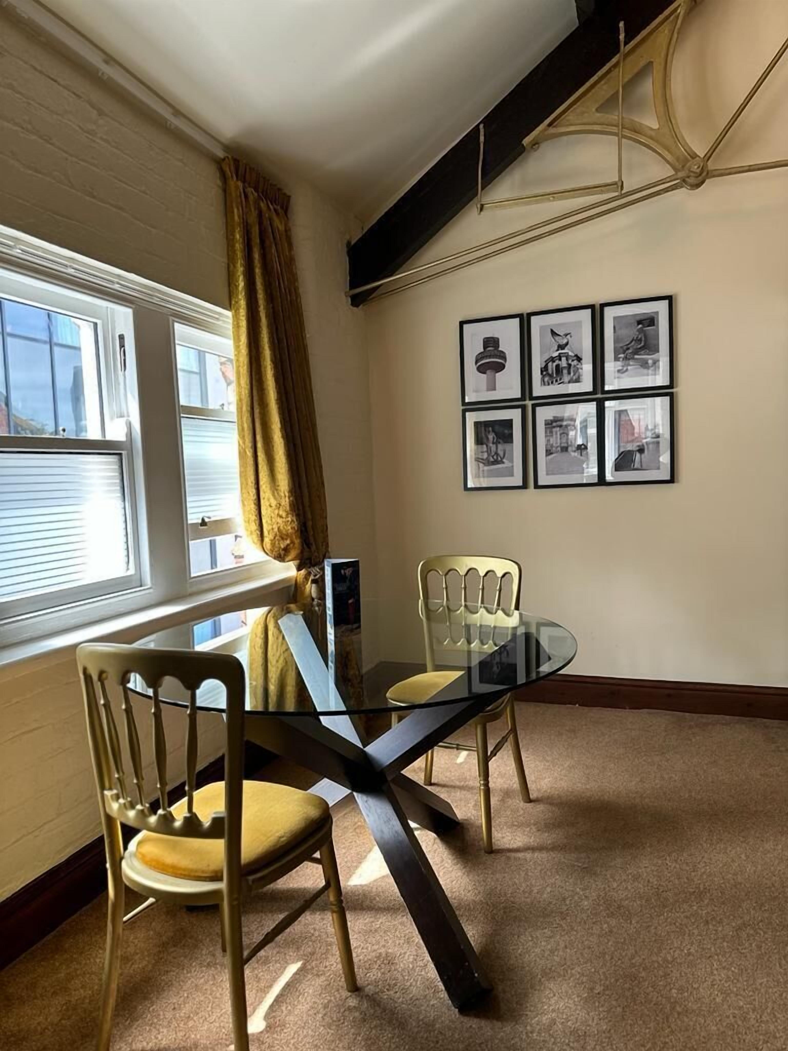 Uma sala de jantar com mesa e cadeiras de vidro, quadros nas paredes e uma janela com cortinas.