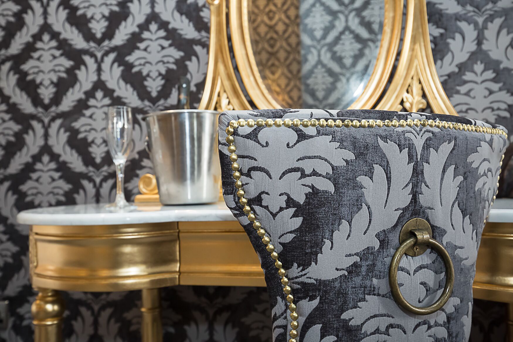 A chair with a patterned fabric and gold bead trim, a metal cup, and a glass on a table with a gold finish.