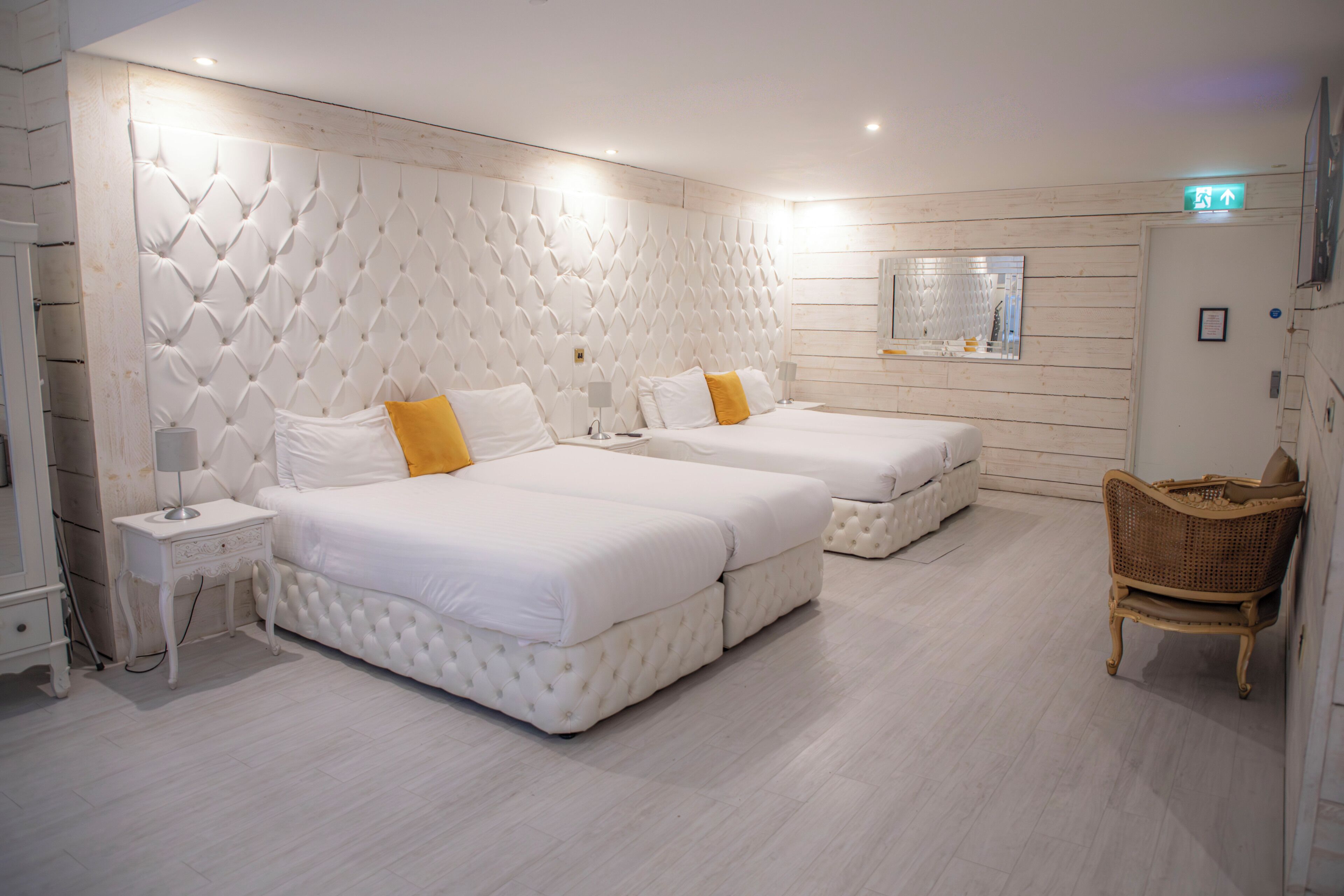 A hotel room with two beds, a small bedside table with a lamp, a wicker chair, and a mirror on the wall.