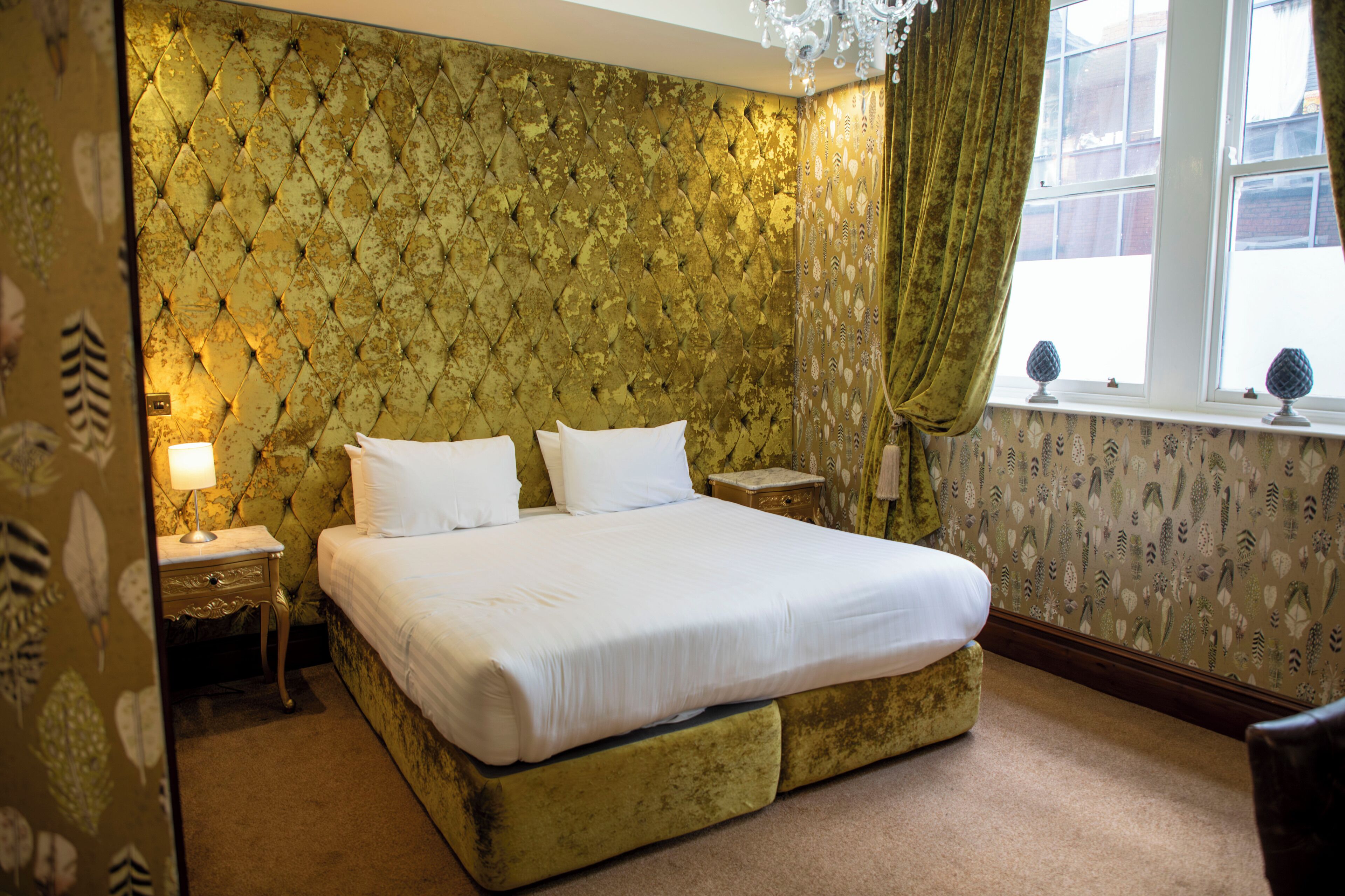 A bedroom with a tufted headboard, a bed with white linens, a nightstand, a lamp, and a window with curtains.