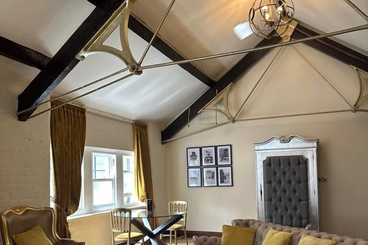 A spacious room with a high ceiling, exposed beams, a tufted headboard, and a large window with curtains.