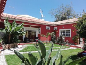 Un edificio rojo con techo de tejas, pared de piedra y jardín con diversas plantas.