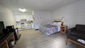 Habitació d'hotel senzilla amb cuina americana, un llit amb llençols estampats florals, una petita tauleta de nit de fusta amb una làmpada i un sofà fosc.