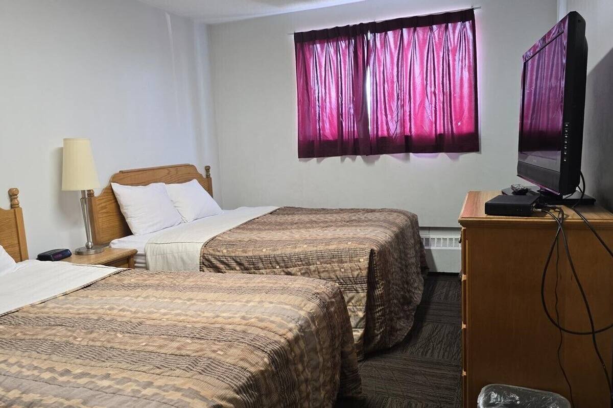 A hotel room with two beds, a television, a desk with a computer, and a window with purple curtains.