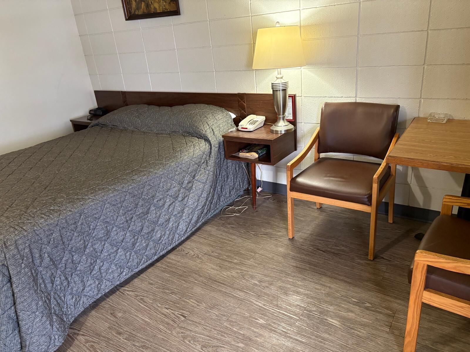 A hotel room with a bed, a desk with a lamp, a chair, and a small table.