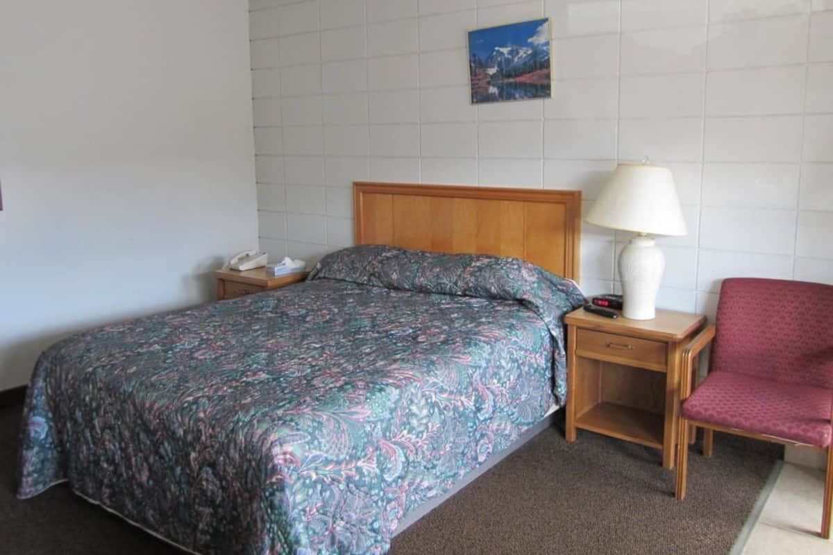 A hotel room with a bed, bedside table, lamp, chair, and a painting on the wall.