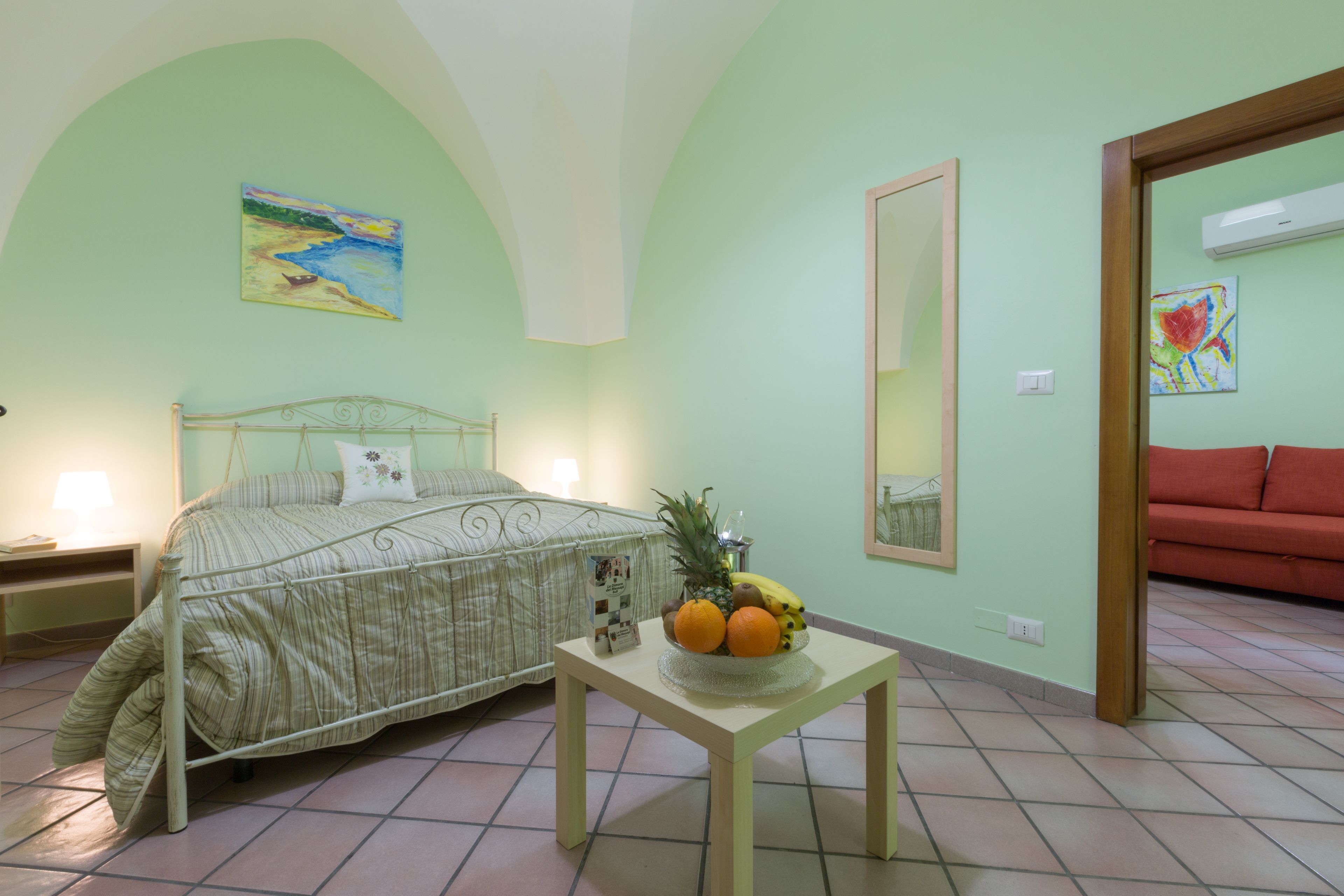 A bedroom with a bed, a table with fruit, a painting, and a mirror.
