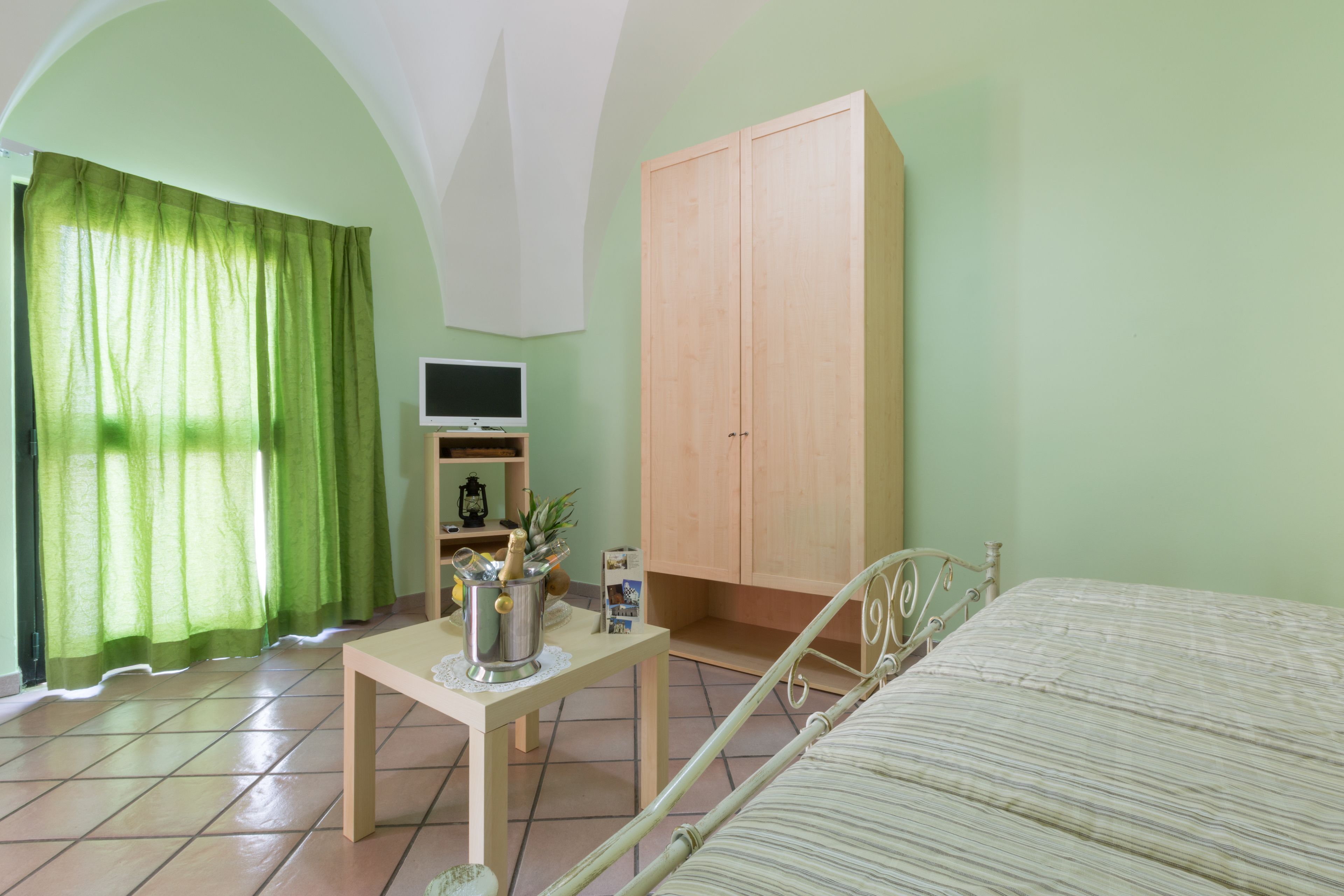 A room with a bed, a wooden cabinet, a television on a stand, and a table with a pitcher and a bottle.