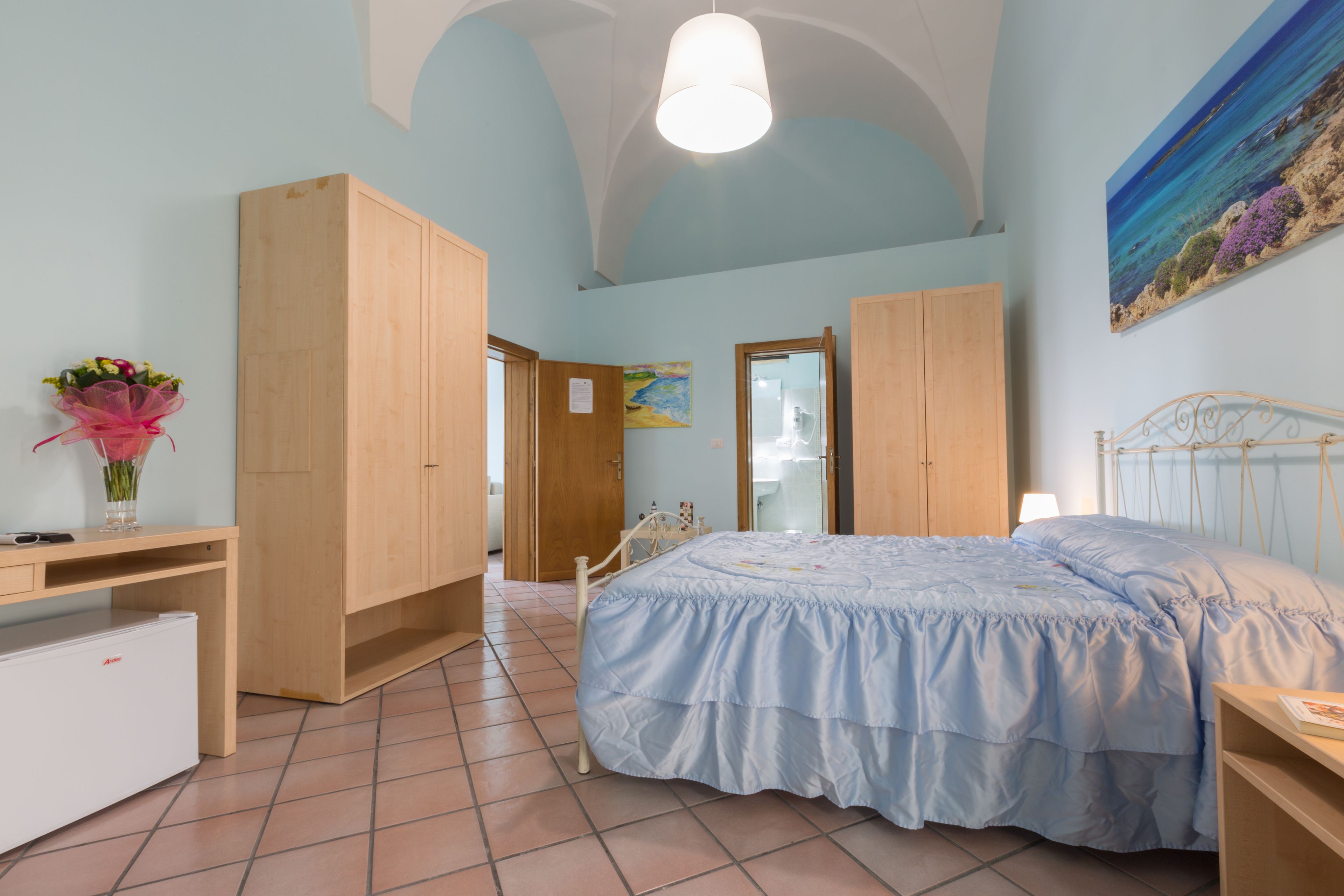 A bedroom with a bed, a desk, a wardrobe, and a small refrigerator.