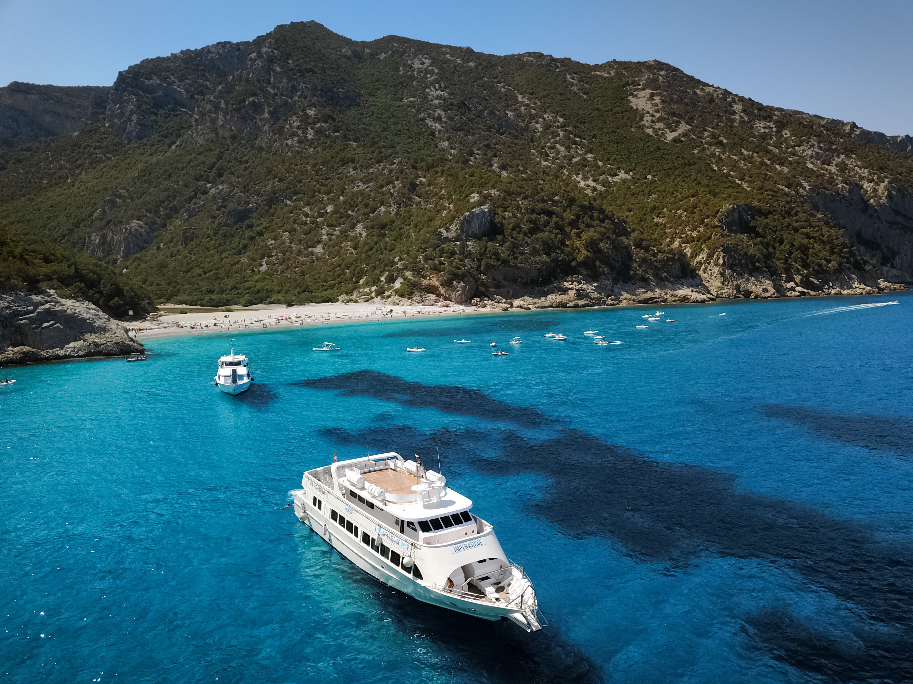 A large white yacht docked at a coastal area with clear turquoise waters, surrounded by rocky cliffs and a sandy beach.