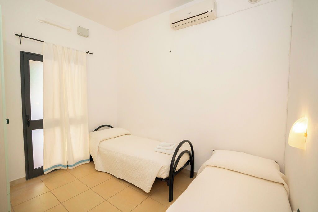 Quarto com duas camas de solteiro, porta com cortina, ar condicionado e luminária fixada na parede.