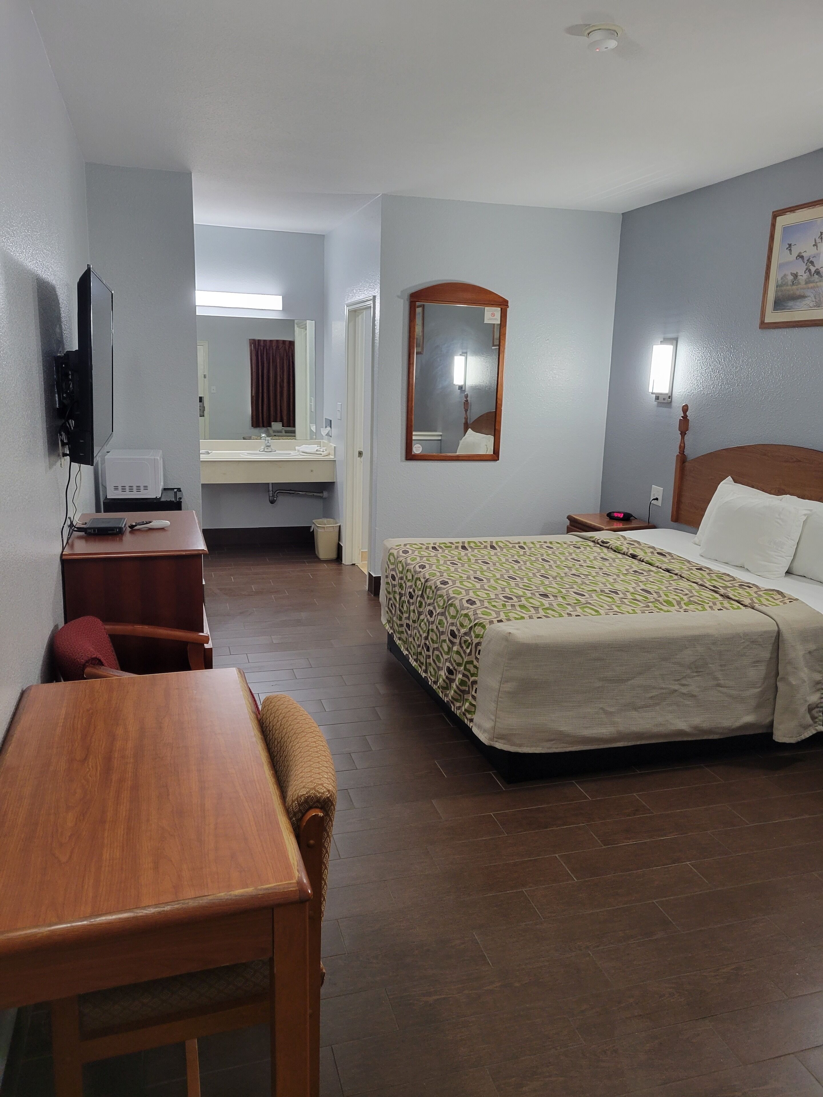 A hotel room with a wooden desk, a bed with a patterned bedspread, a mirror, and a television on the wall.