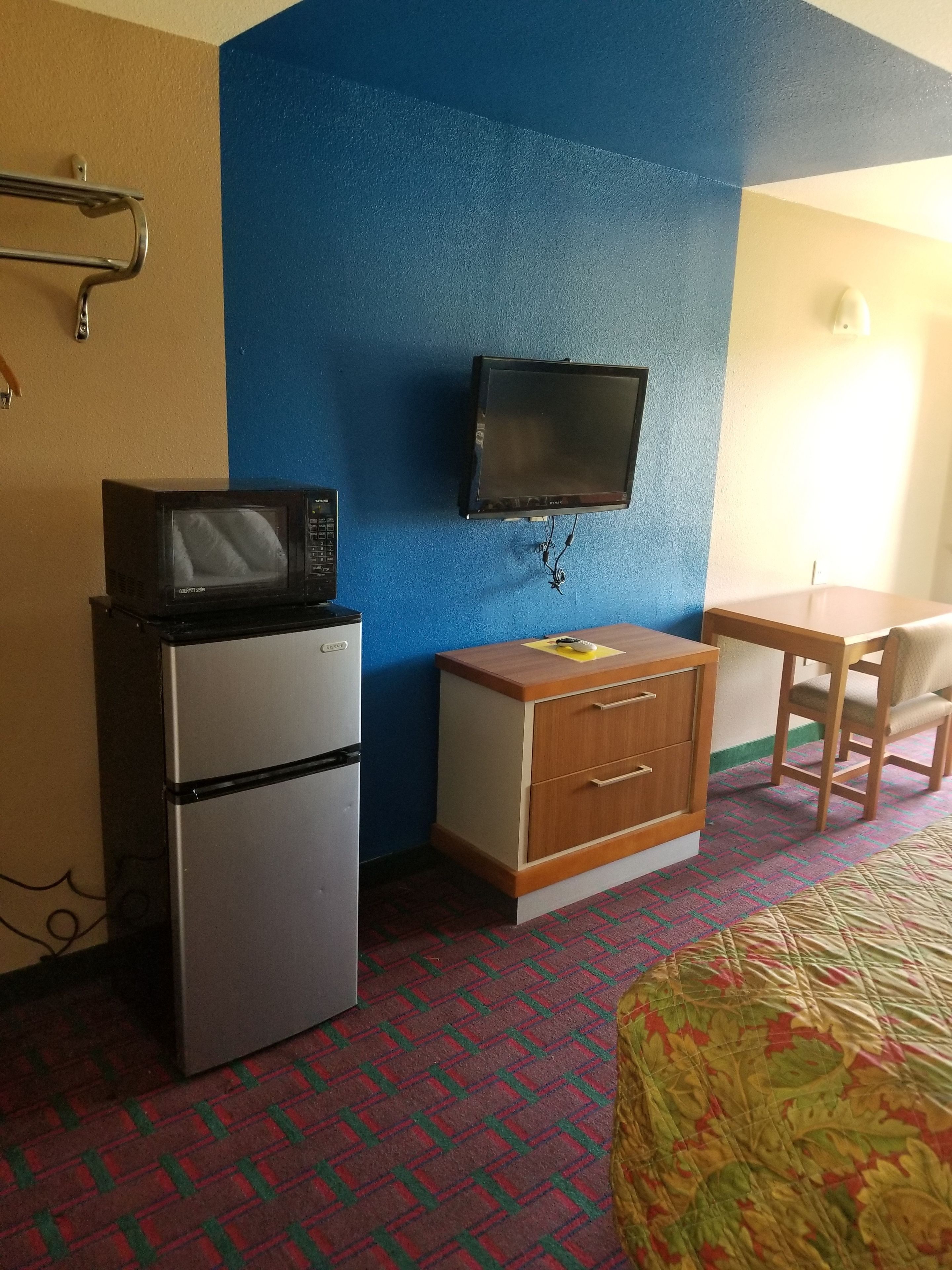 A small, compact room with a kitchenette, a television mounted on the wall, a bed with a patterned cover, and a chair.