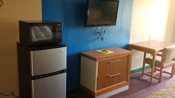 A small, compact room with a kitchenette, a television mounted on the wall, a bed with a patterned cover, and a chair.