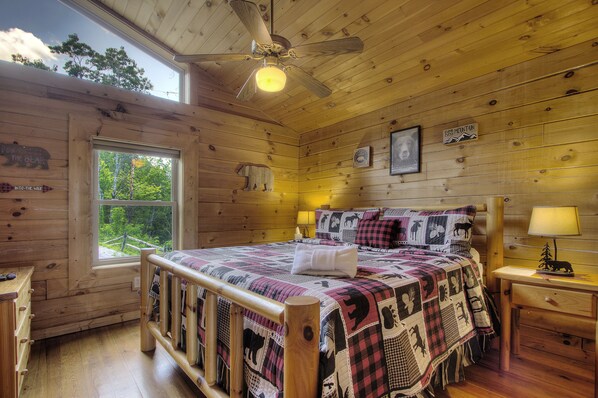Un dormitorio de cabaña de madera con cama, mesita de noche, ventilador de techo y ventana con vista a árboles.
