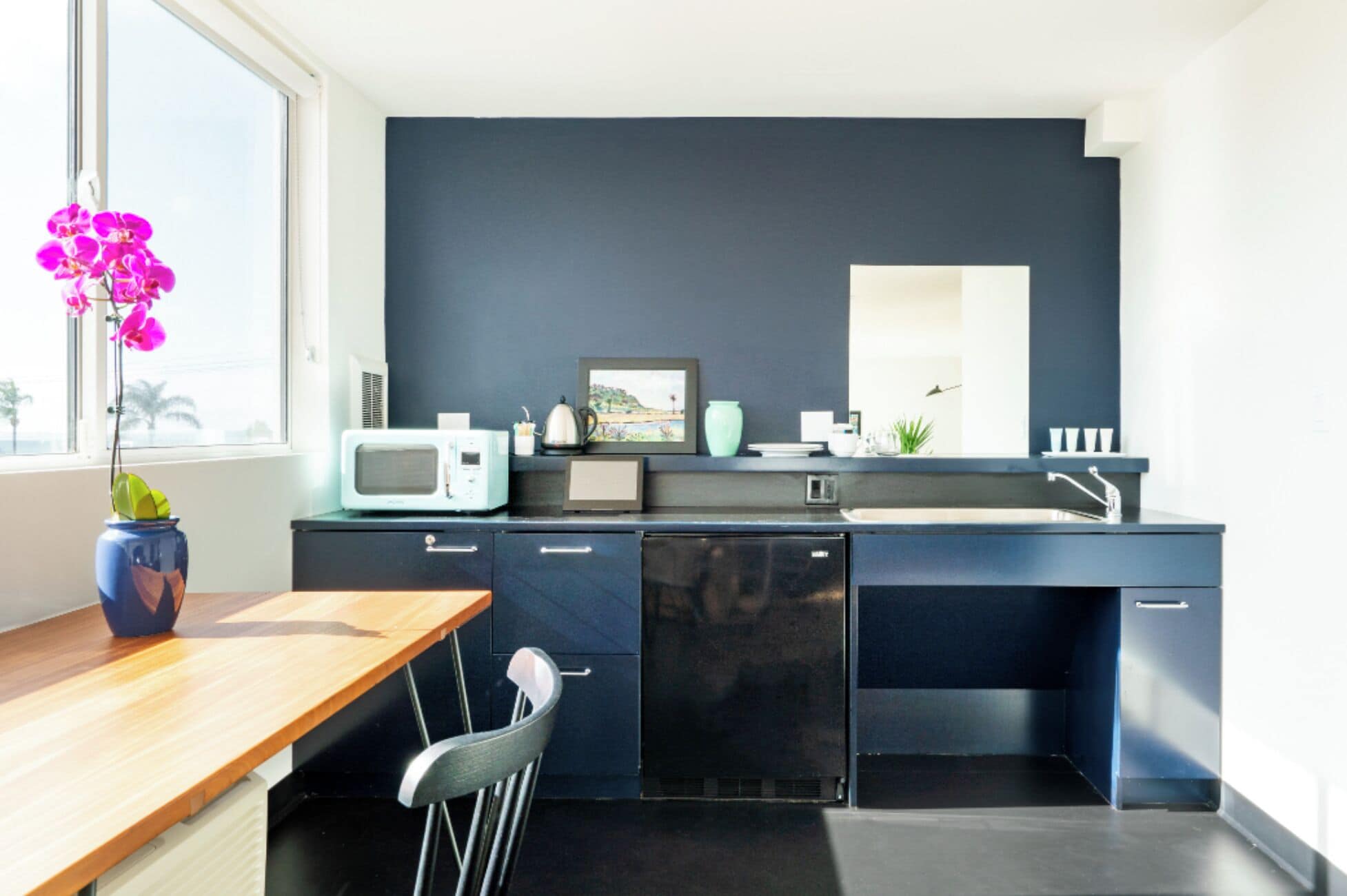 Una cocina moderna con pared azul oscuro, microondas, fregadero y ventana con vista a palmeras.