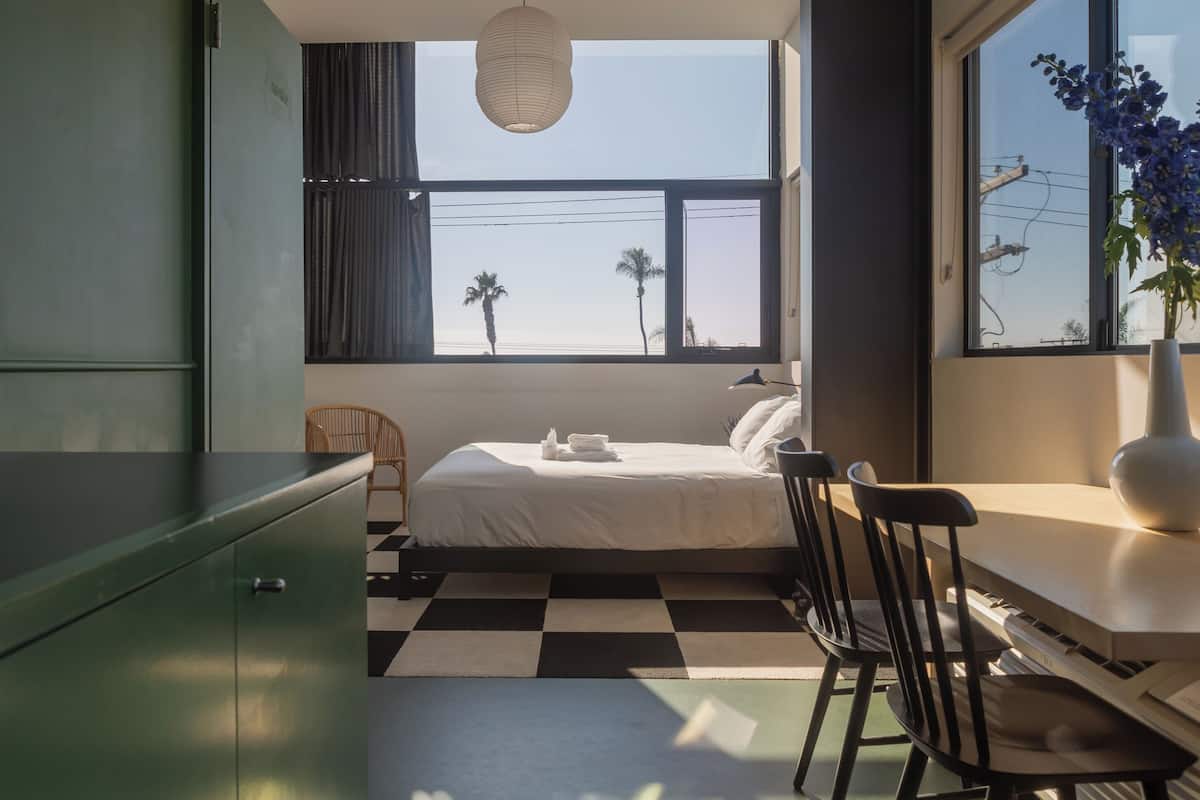 Quarto de hotel moderno com cama, piso xadrez, escrivaninha com teclado, cadeira, luminária e janela com vista para palmeiras.