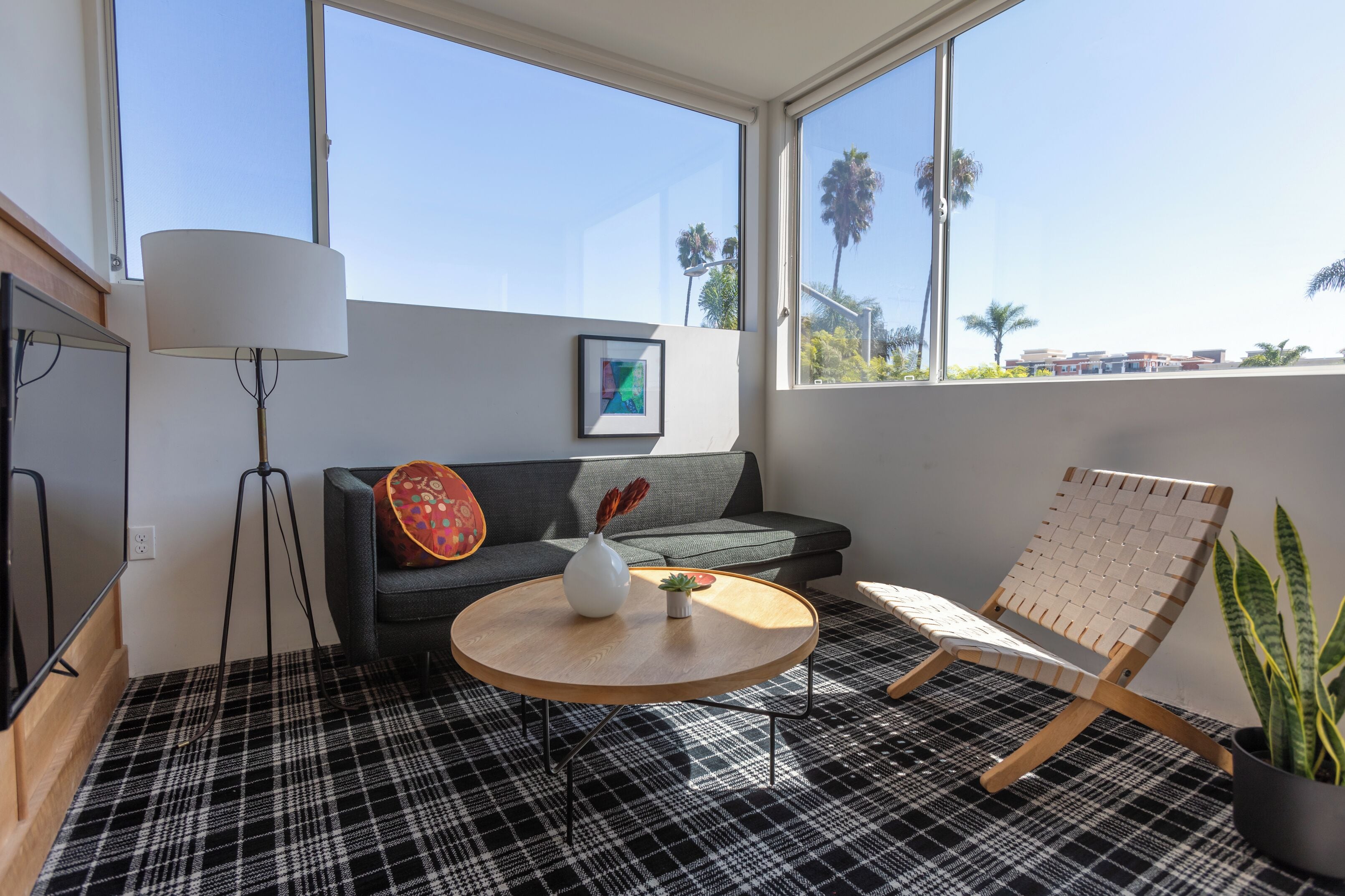 A modern living room with a sofa, a round coffee table, a floor lamp, and a view of palm trees through large windows.