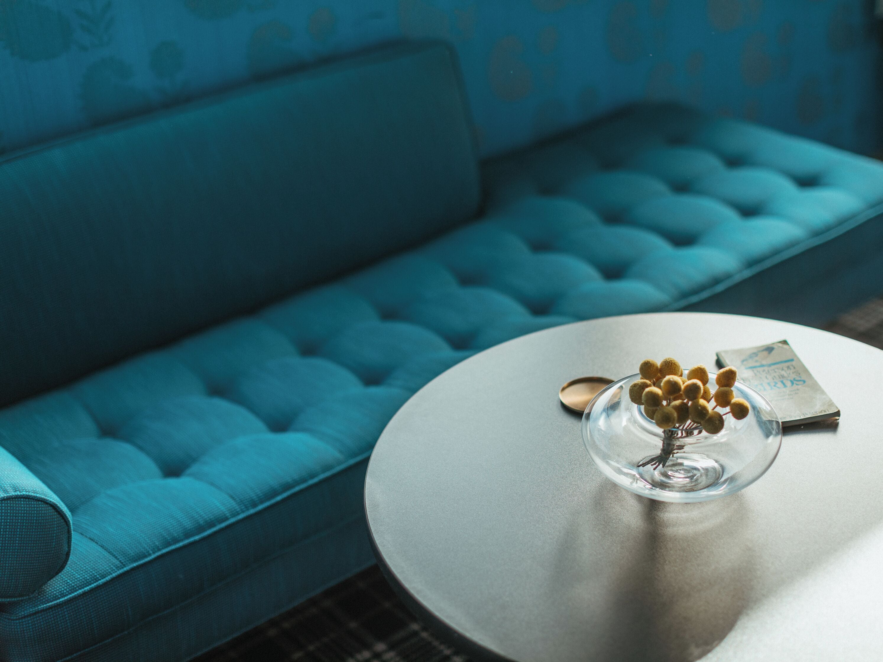 A blue sofa with a tufted design and a round table with a glass bowl containing small items and a book.