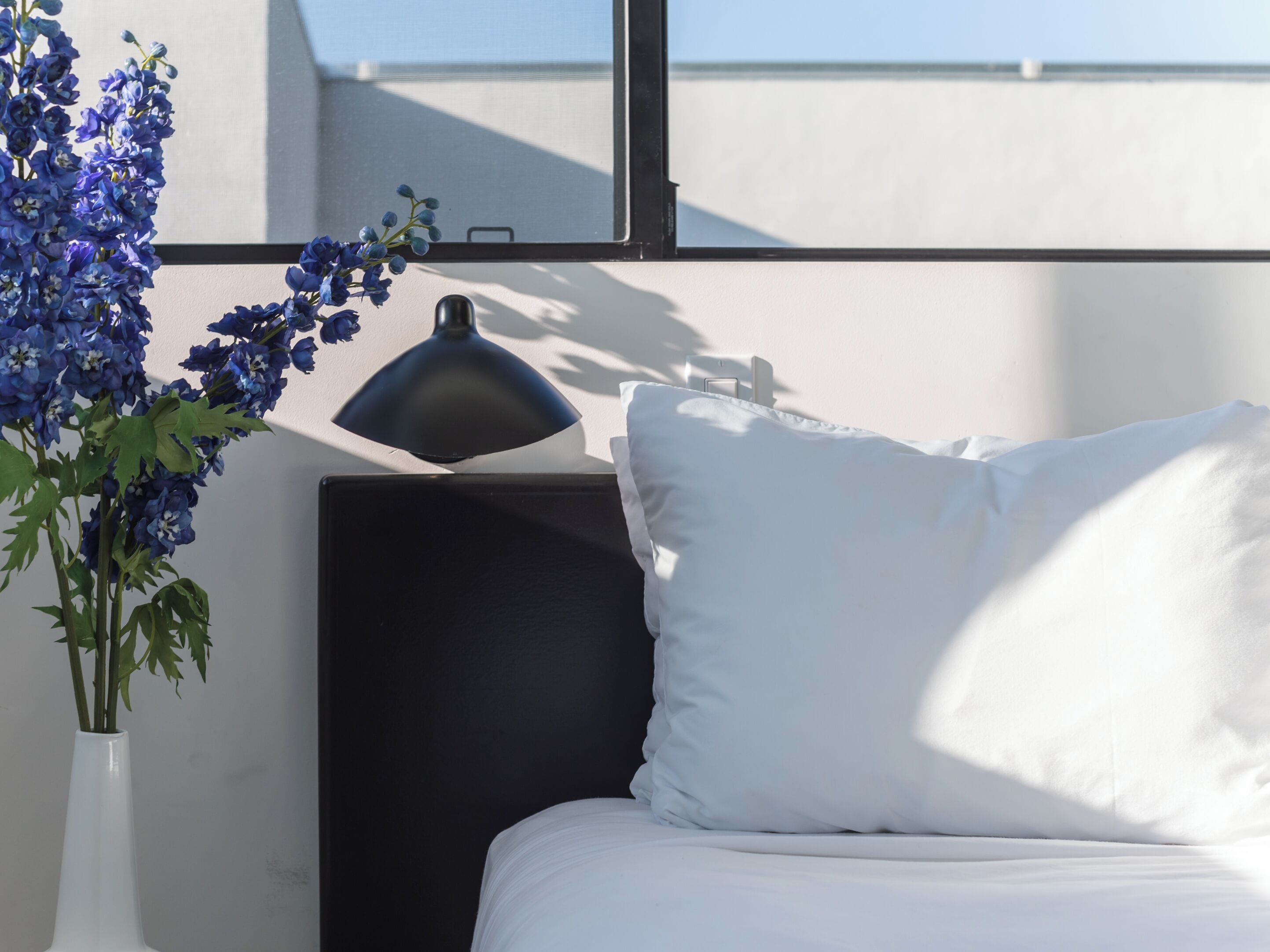 A bed with white bedding, a black headboard, and a white pillow. There is a black desk lamp on the headboard. A vase with blue flowers is visible on the left side of the image.