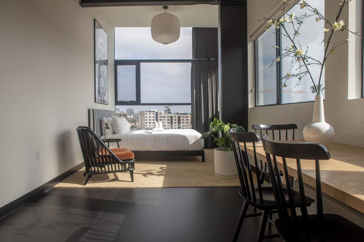 Quarto moderno com uma cama grande, uma mesa de jantar com cadeiras, uma planta em vaso e vista para prédios através de grandes janelas.