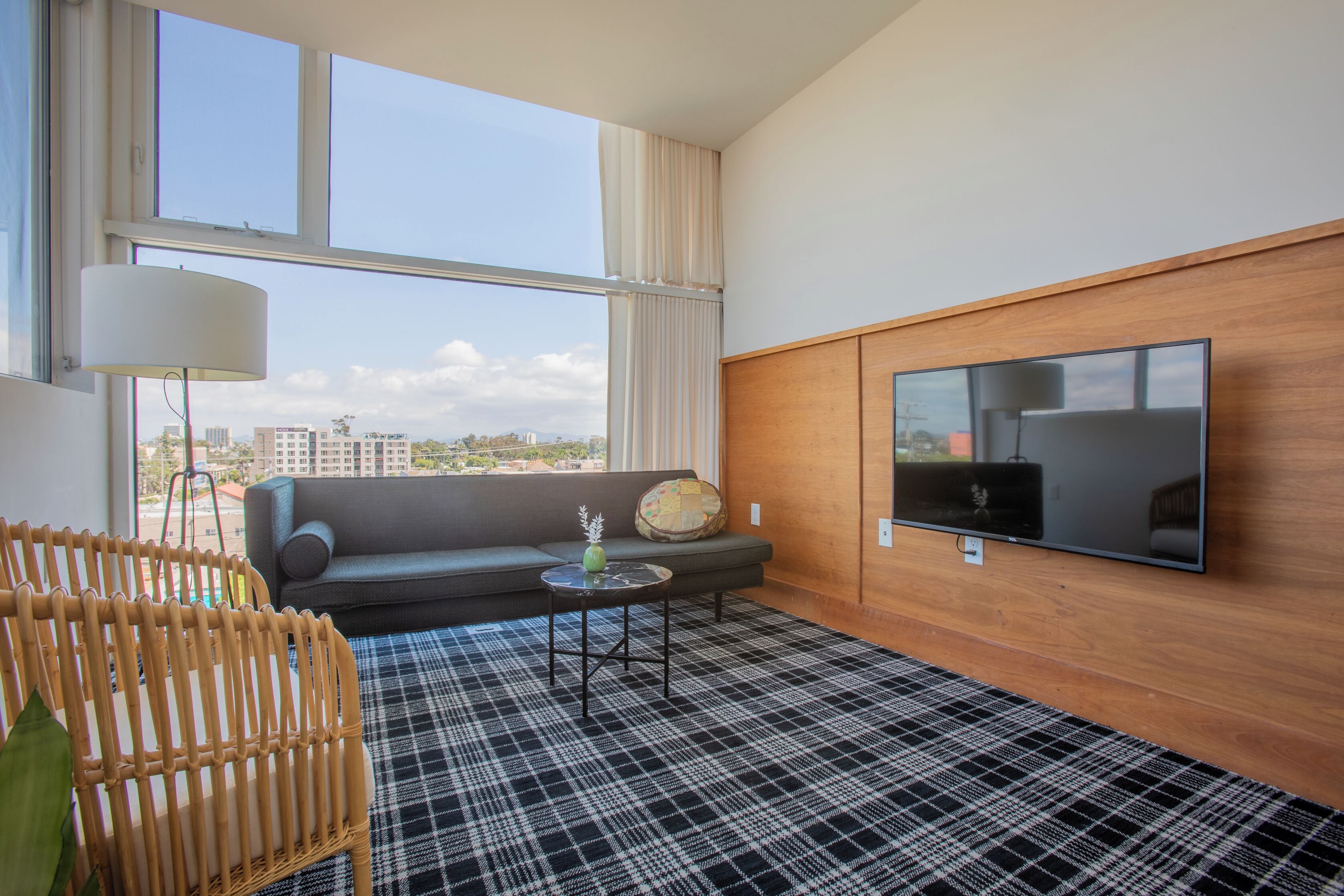 A modern hotel room with a large window, a flat-screen TV, a sofa, a small table, and a checkered floor.