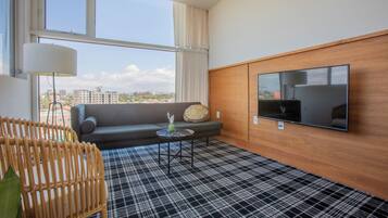 A modern hotel room with a large window, a flat-screen TV, a sofa, a small table, and a checkered floor.