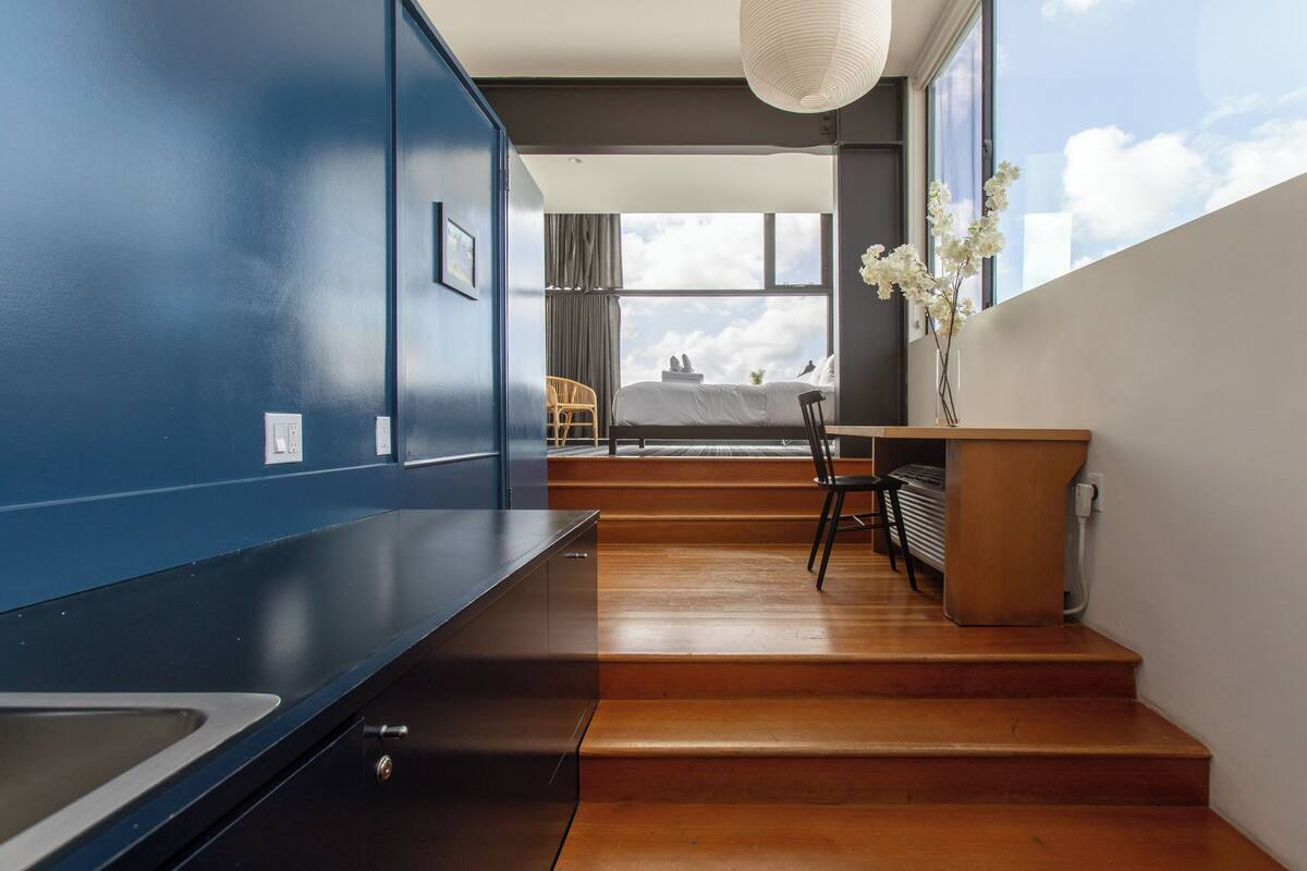 Uma cozinha moderna com bancada de madeira, pia e vista para um quarto com cama e cadeira.