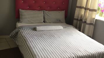 Ein Bett mit gestreifter Bettwäsche, ein roter, gepolsterter Kopfseitenverkleidung und ein Fenster mit Vorhängen.