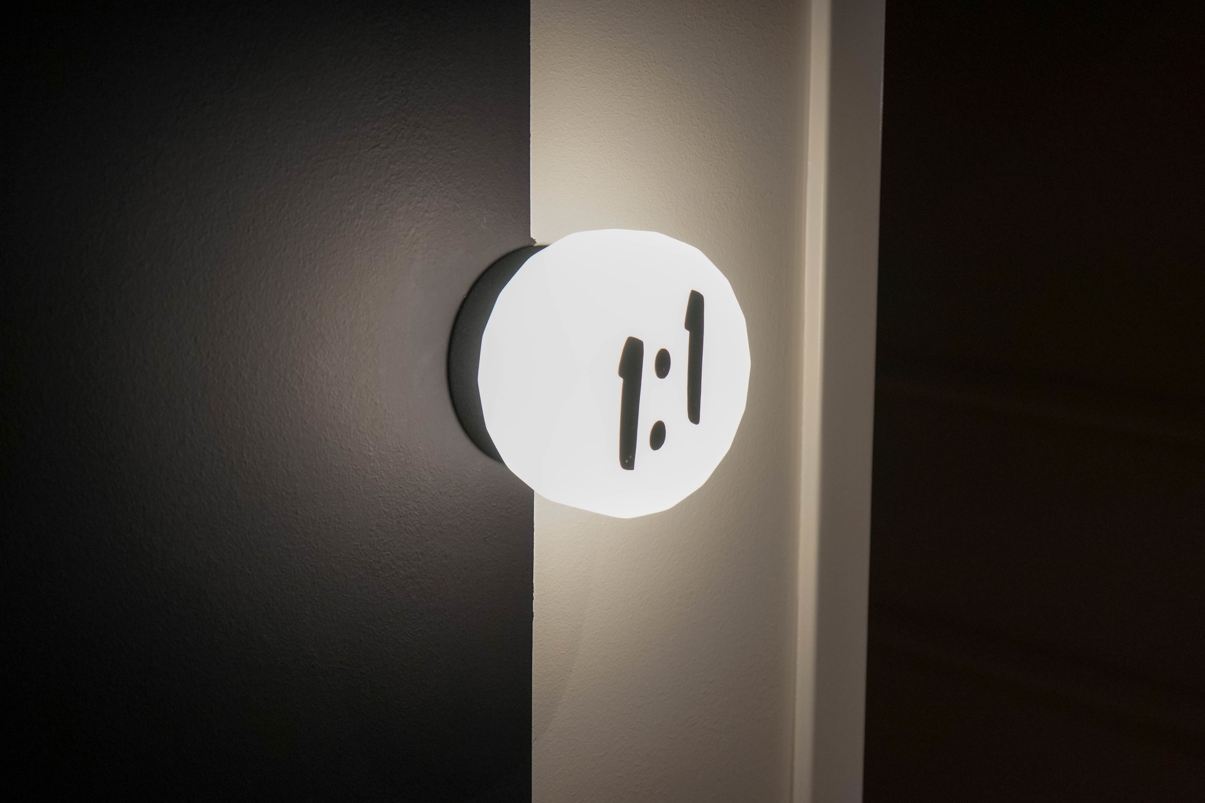 A round, illuminated sign with a colon and a vertical line, mounted on a wall.