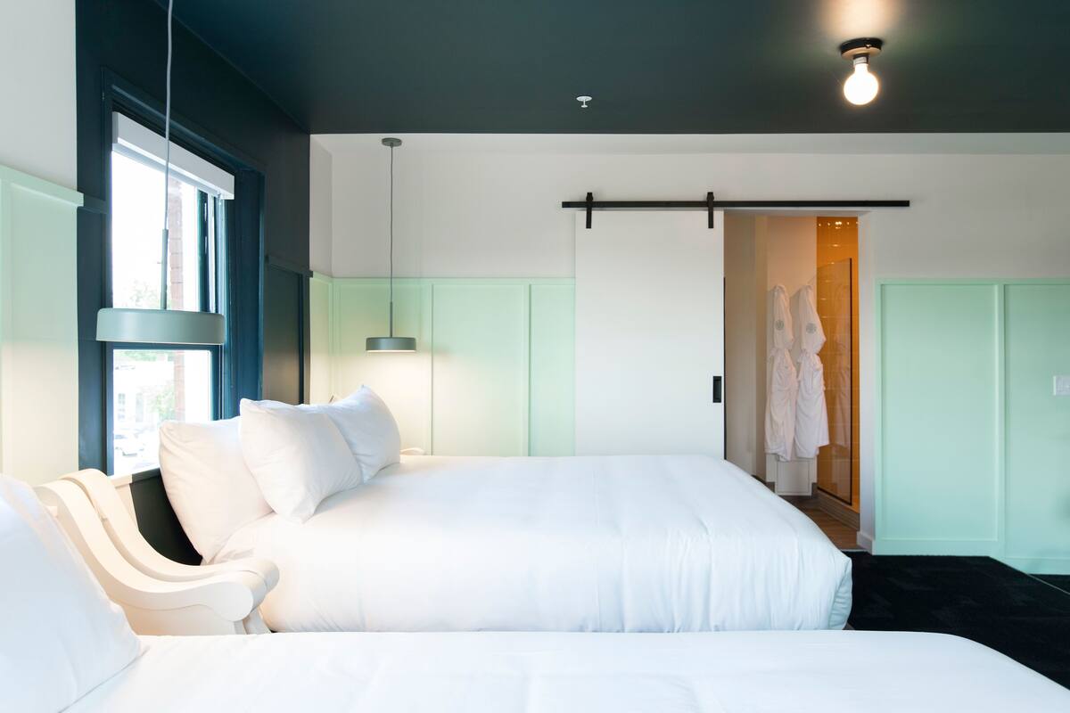 Ein modernes Hotelzimmer mit einem großen Bett, einem Nachttisch, einer Hängelampe und Blick ins Freie durch ein Fenster.