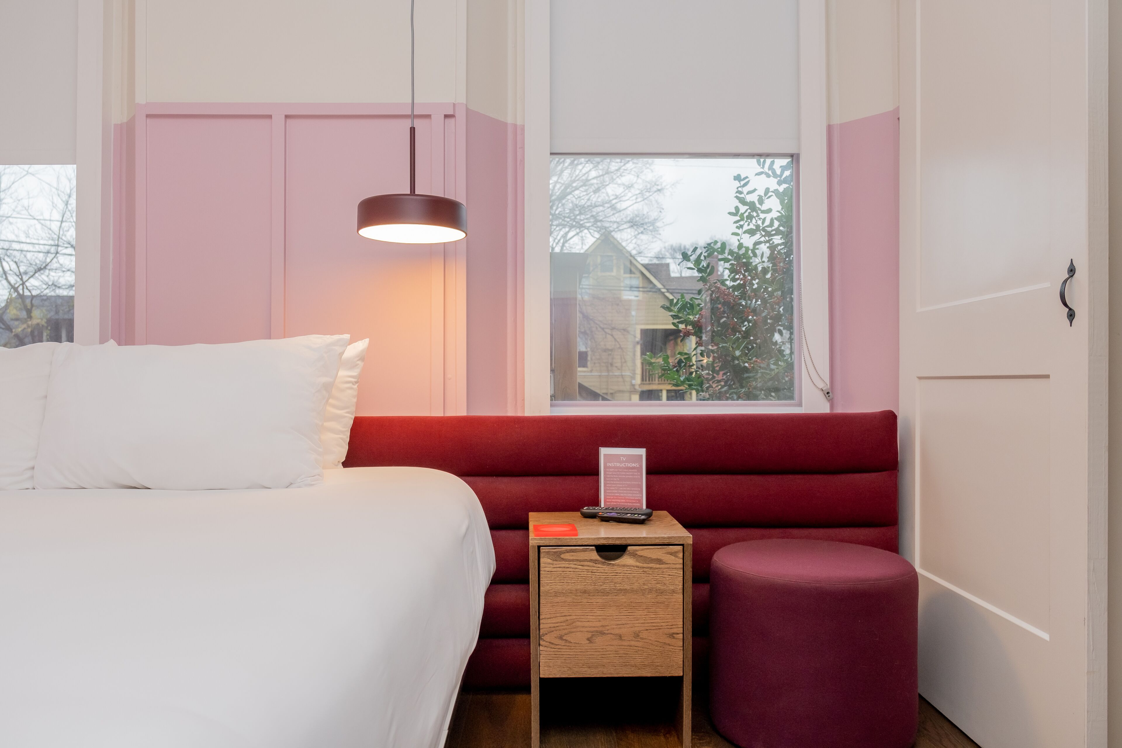 A bedroom with a bed, a bedside table with a lamp, a red upholstered bench, and a window with a view of houses.
