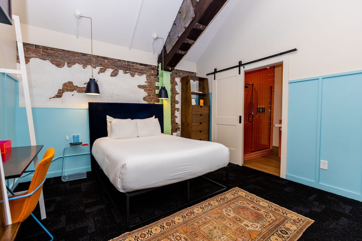 Ein Schlafzimmer mit Bett, Schreibtisch, Stuhl, Teppich und einem durchgehenden Türrahmen sichtbaren Badezimmer.