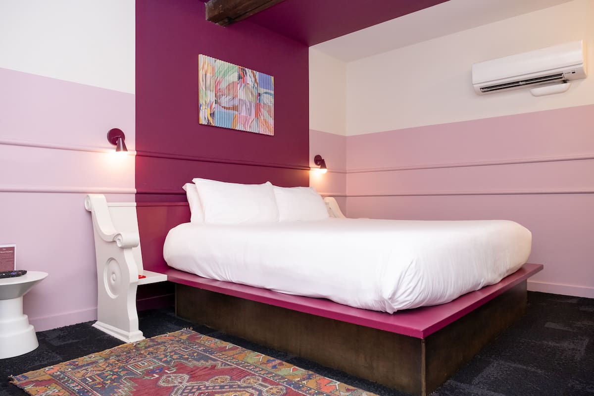 Ein Schlafzimmer mit einem Bett, einem Nachttisch, einem Stuhl, einem farbenfrohen Teppich und einer Klimaanlage.