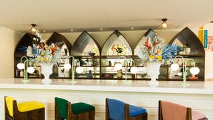 Una zona de bar con una barra blanca, varios taburetes con cojines de colores y jarrones decorativos con flores.