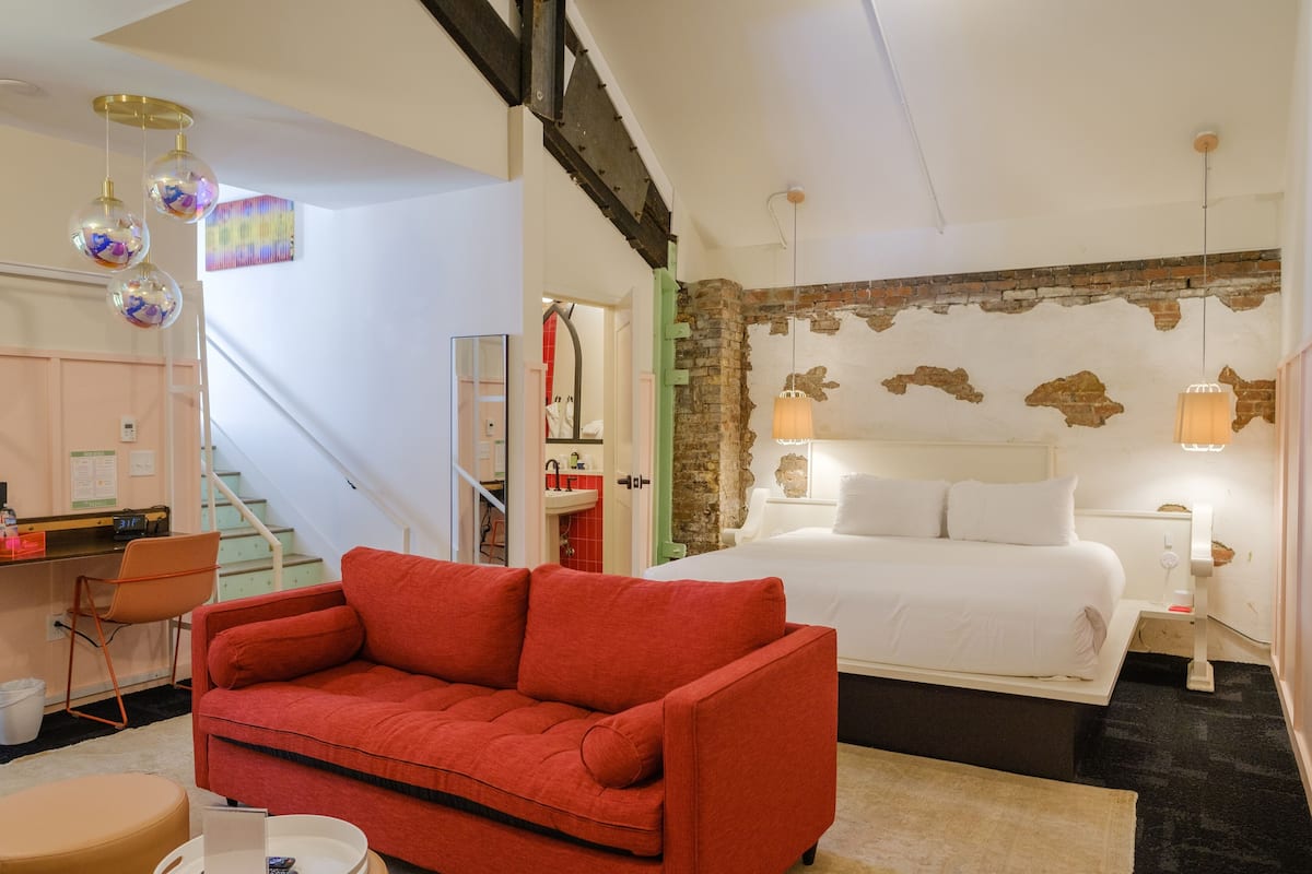 Ein Hotelzimmer mit rotem Sofa, einem Bett mit weißer Bettwäsche, einer Wand aus Backsteinen und einer Treppe, die in einen anderen Raum führt.